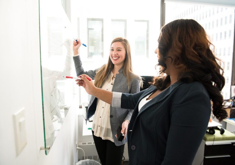 Two women writing on a whiteboard, smiling.