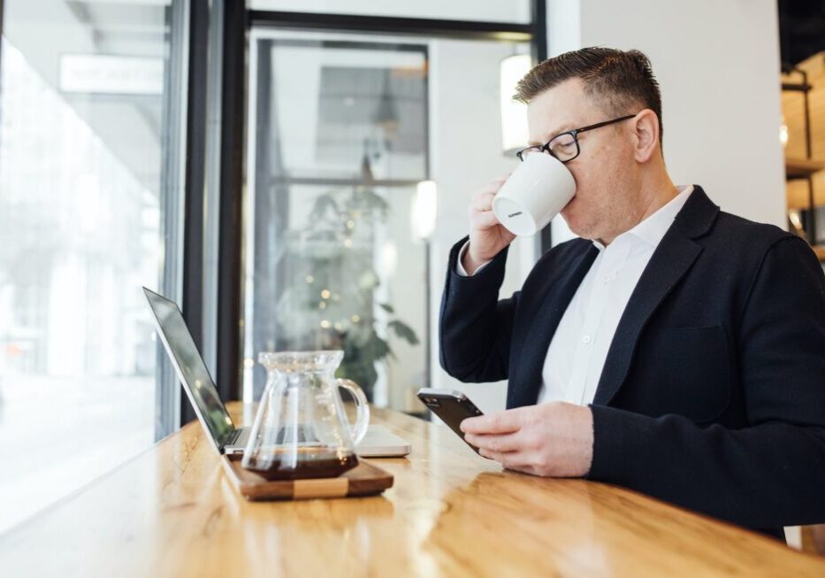Man drinking coffee, using phone and laptop.