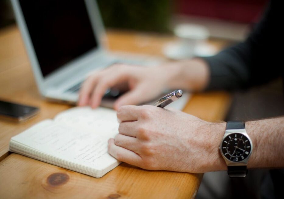 Person writing in notebook beside laptop.